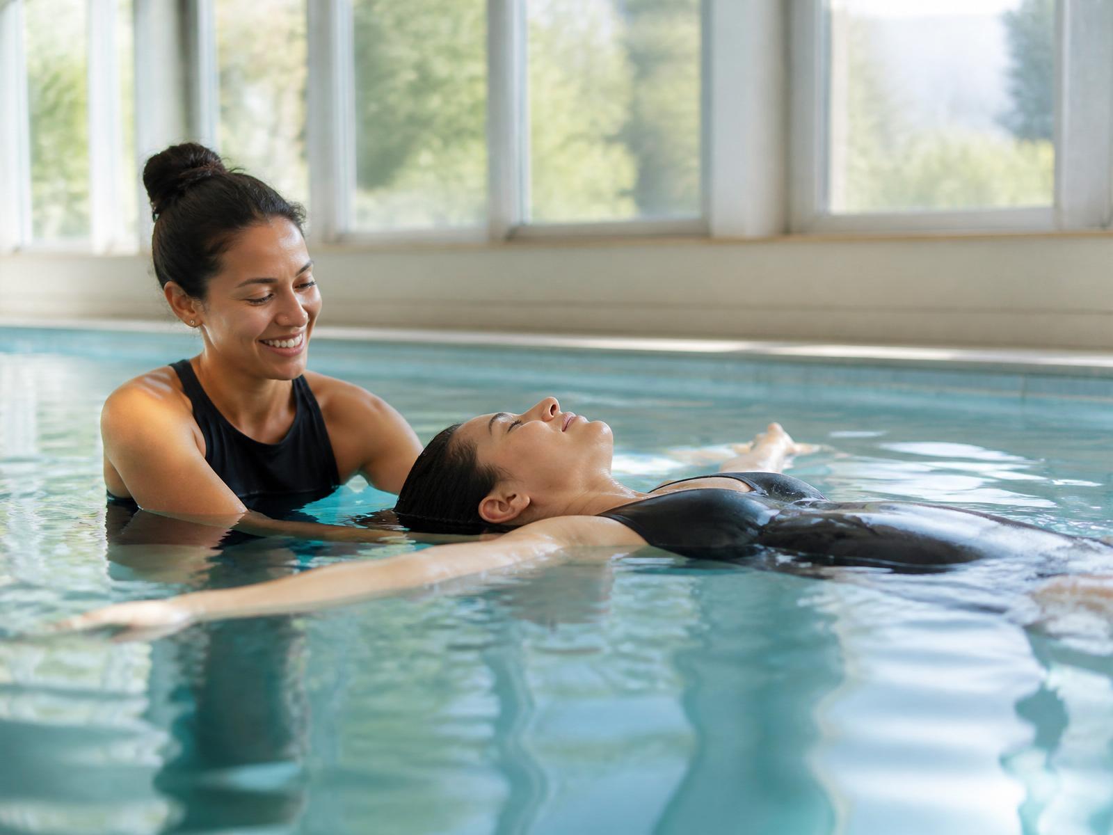 A swim instructor calmly supporting an adult learner practicing a back float in a bright indoor pool.